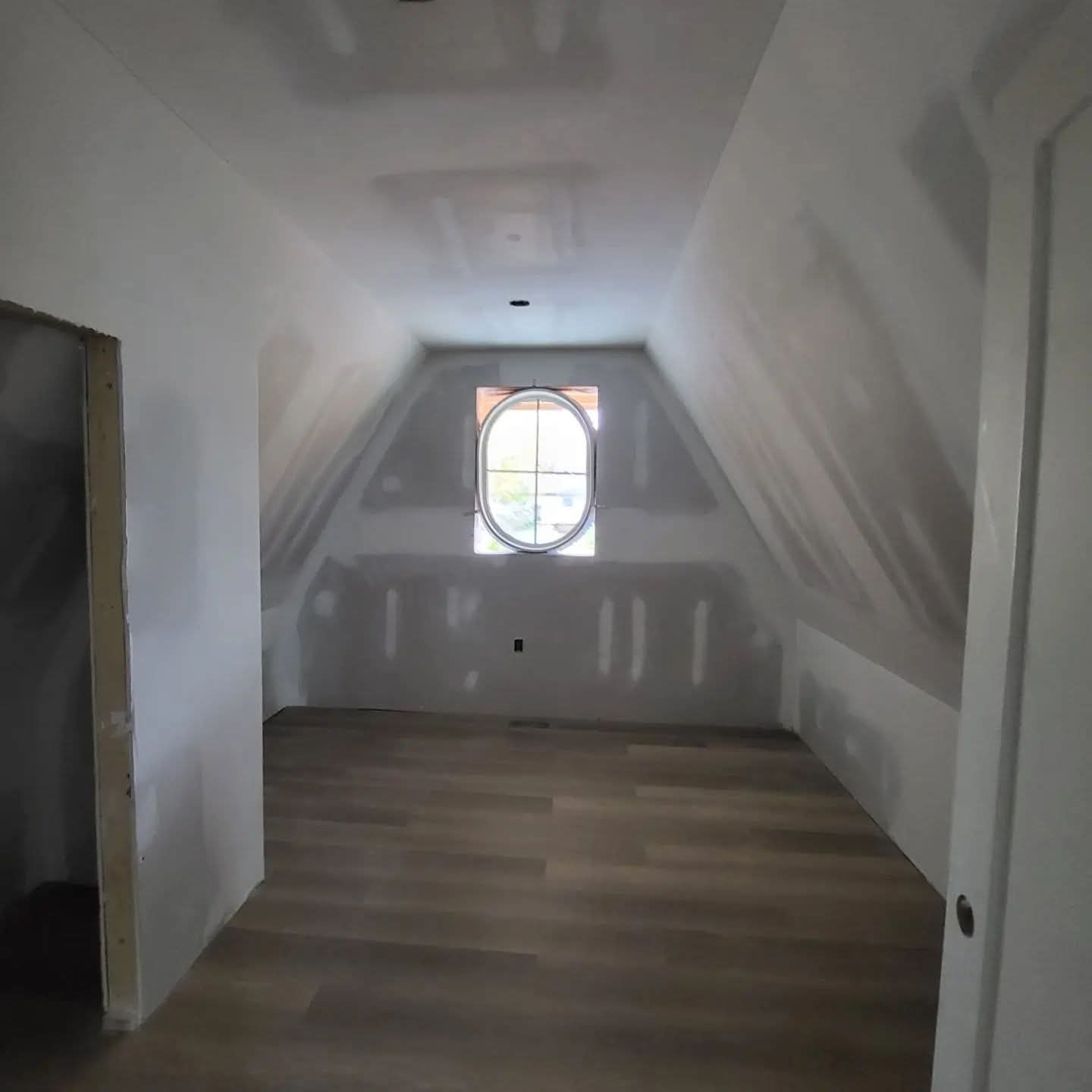 Finished upper-floor room with sloped drywall walls, smooth ceiling, and oval window
